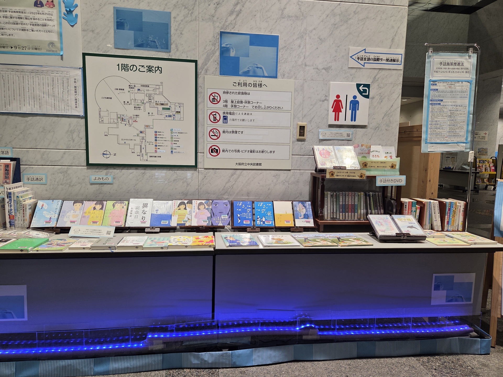 資料展示「手話の日・手話言語の国際デーを知っていますか?」の展示風景写真。机の上に表紙を見せた本やDVDが並んでいて、机の下からは青いライトで照らされている。