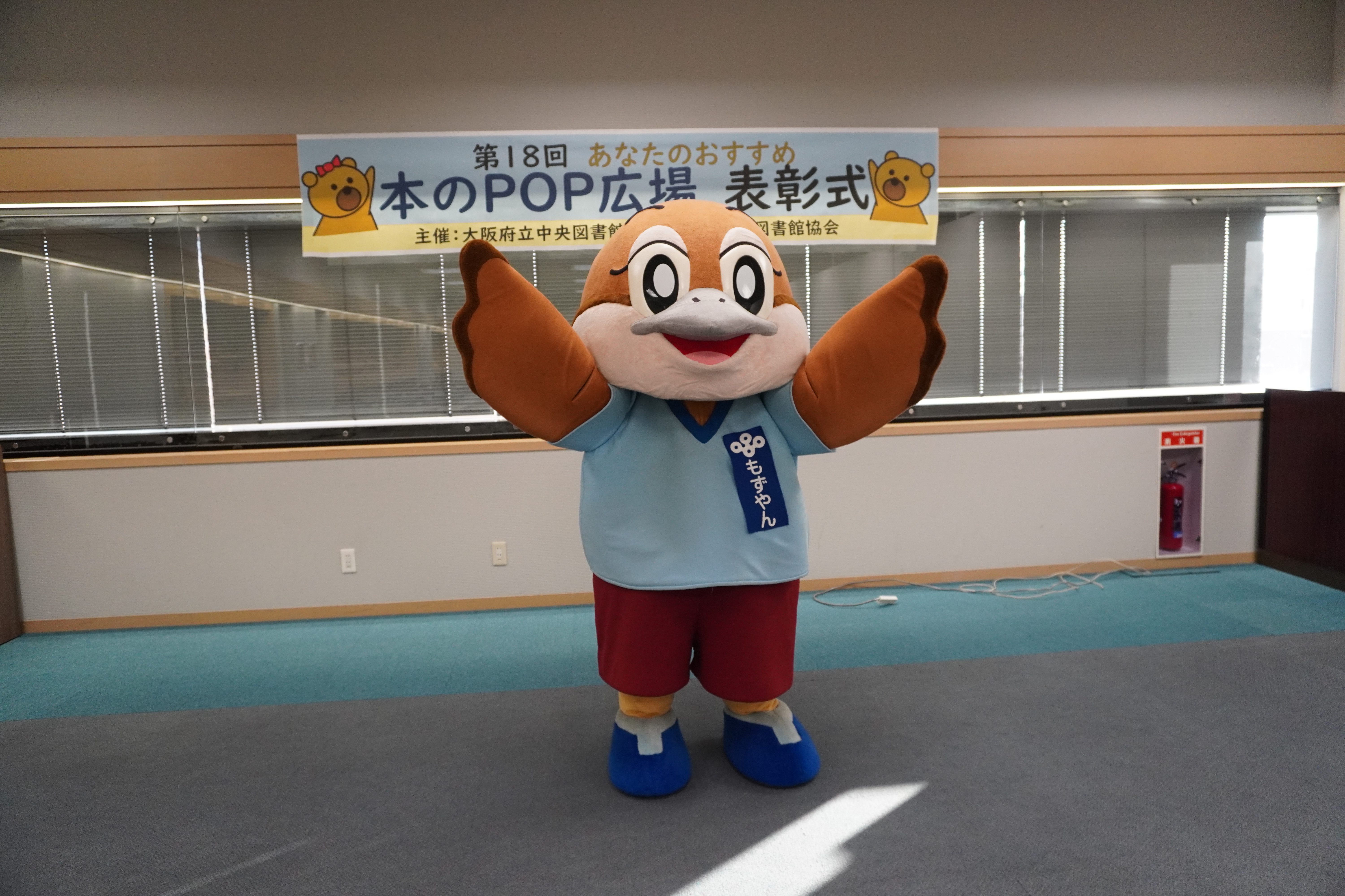 大阪府の広報担当副知事もずやんが表彰式でお祝いしている様子。表彰式と書かれた幕の前でポーズをとっている。