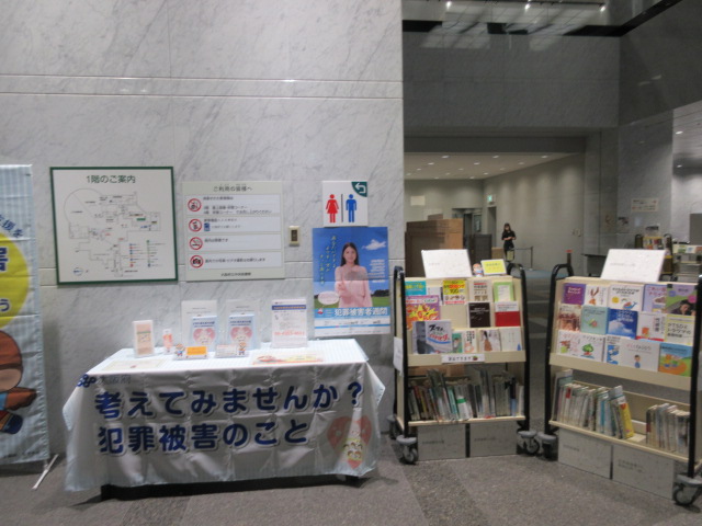 昨年度の展示の様子 机には「考えてみませんか?犯罪被害のこと」と書かれた布がかけらている。その右側には貸出可能な展示に関連する図書がブックトラックにのせられている。