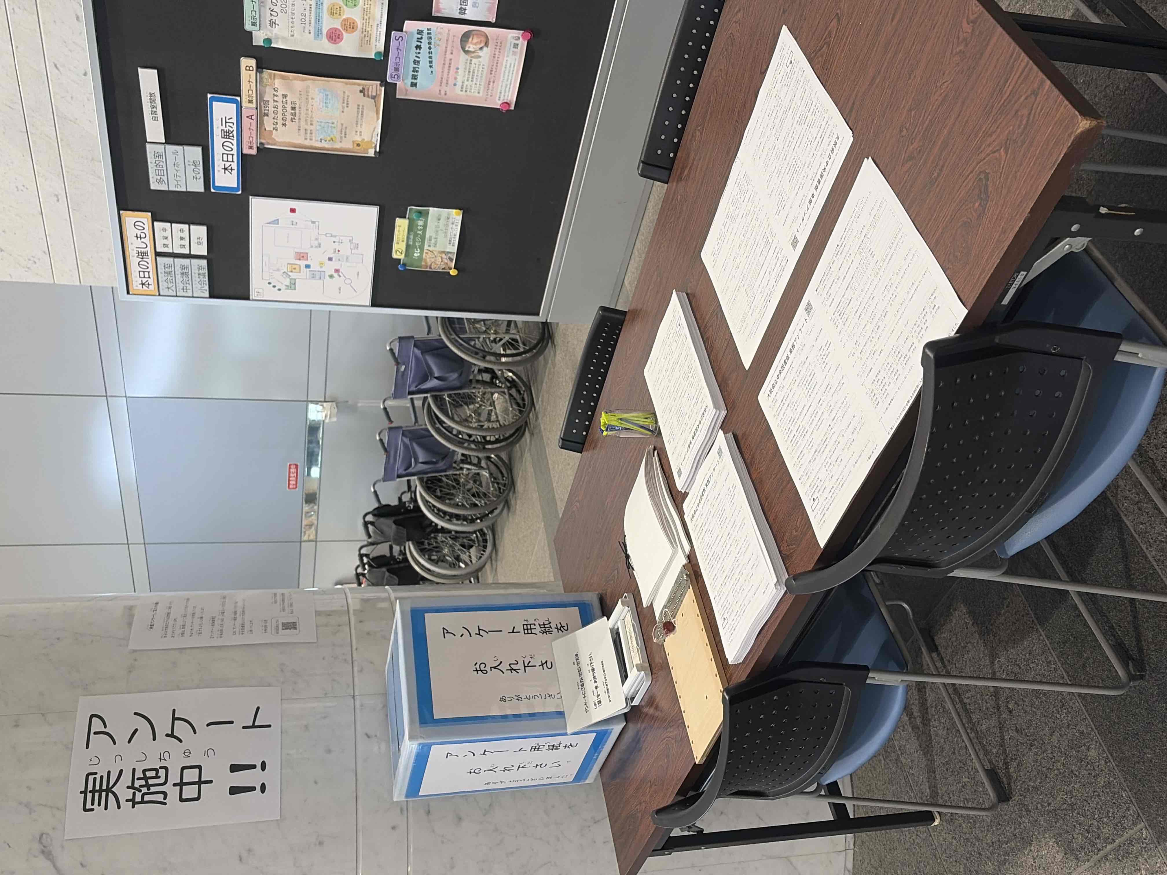 エントランスの柱の前に設置されたアンケート記入台と回収箱の様子。記入台の手前には墨字版のアンケート、奥には点字版のアンケートと点字器が置かれている。