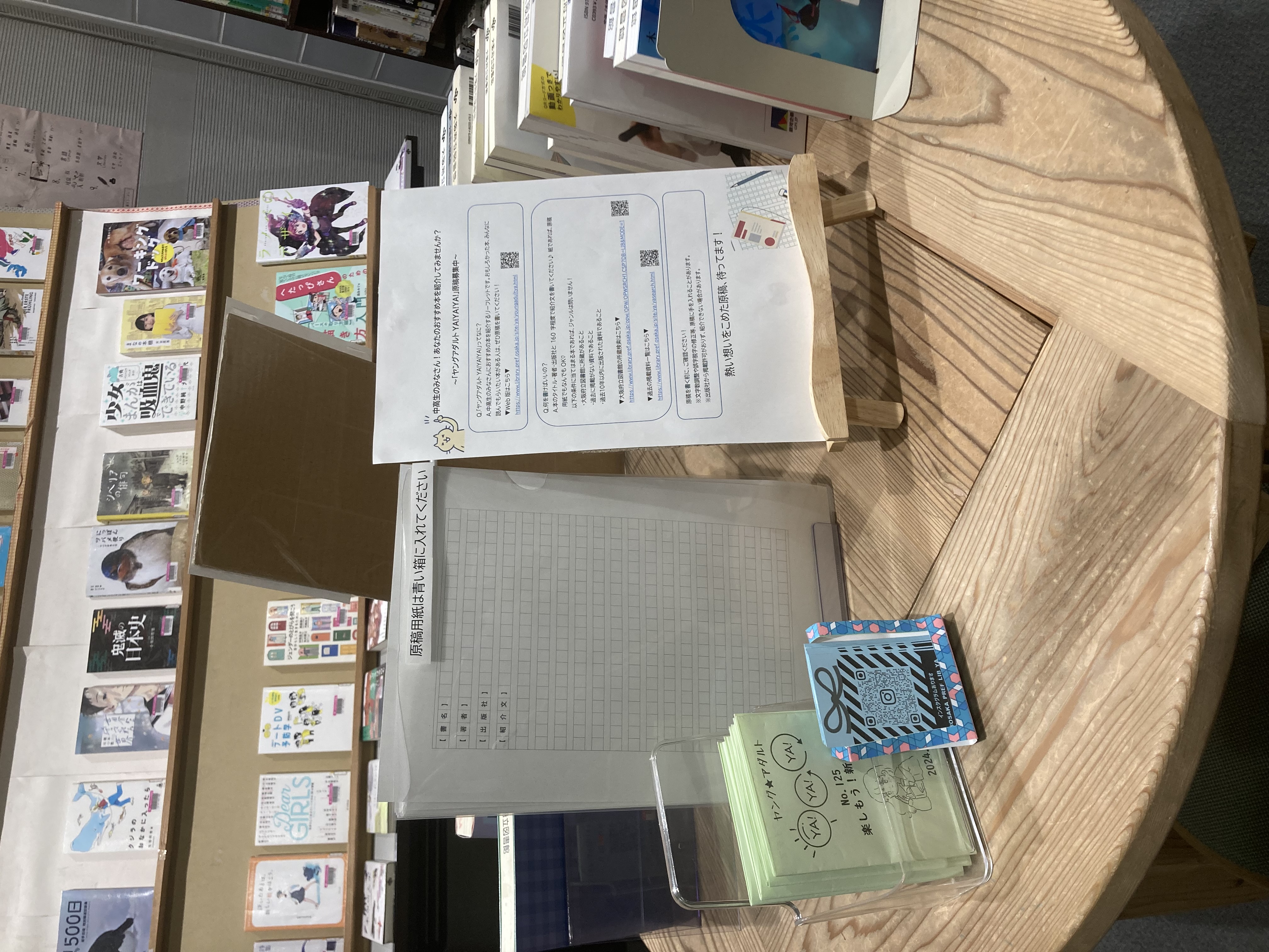 机の上に、ヤングアダルトYA!YA!YA!原稿募集箱と原稿用紙、原稿募集に関するチラシが展示してある風景の写真。原稿募集箱の横には、リーフレット「ヤングアダルト YA!YA!YA!」も置いてある。