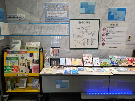 資料展示「手話言語の国際デーを知っていますか?」の展示風景写真。展示見出しの下に年表、その下、左手には本をのせる展示台に表紙を見せて本が並んでいる。その右側に机の上にも表紙を見せた本やDVDが並んでいる。机の下からは青いライトで照らされている。