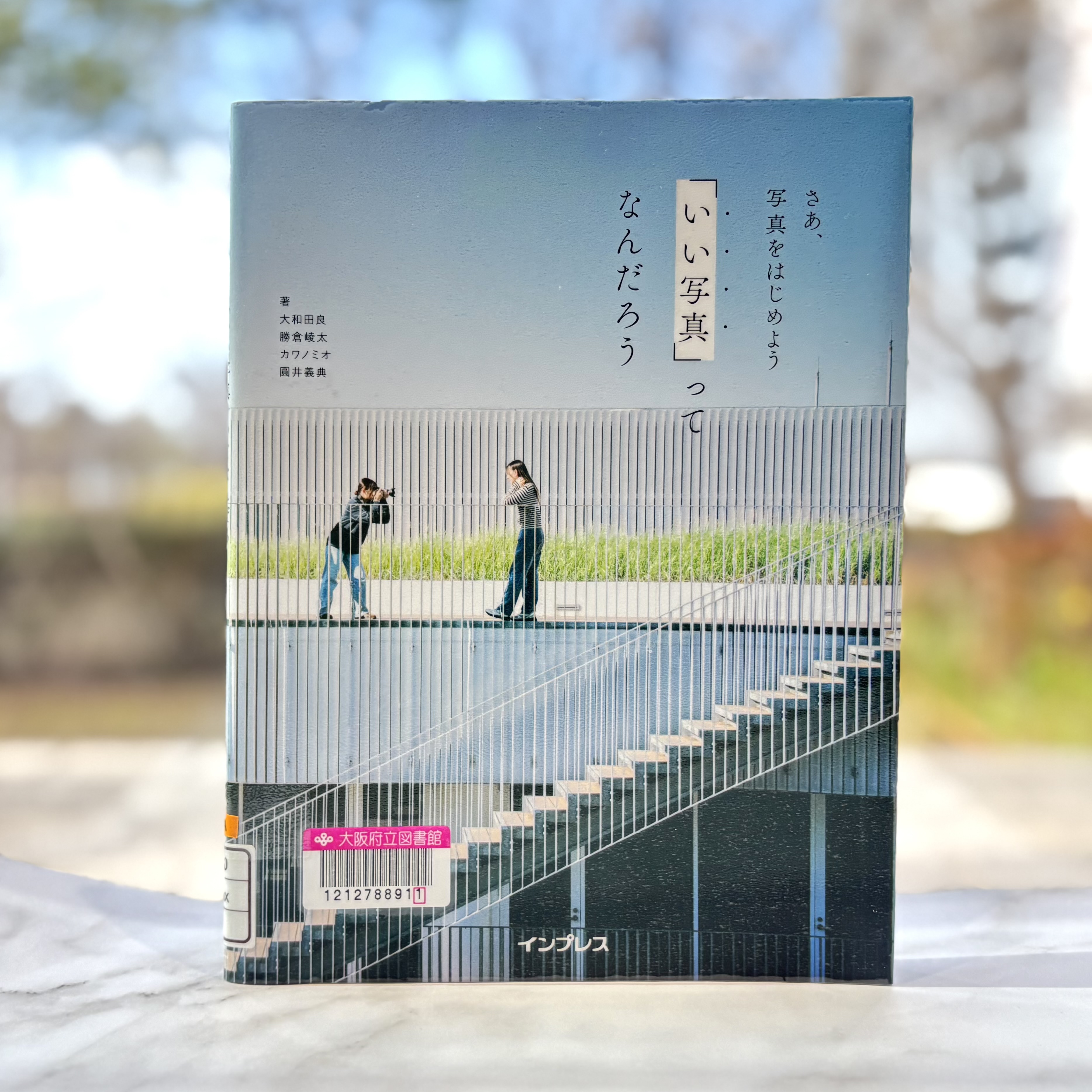 『「いい写真」ってなんだろう：さあ、写真をはじめよう』の表紙画像。青空の下、近代的な建物の階段付近でカメラを構える人物とポーズをとる人物が描かれた表紙。