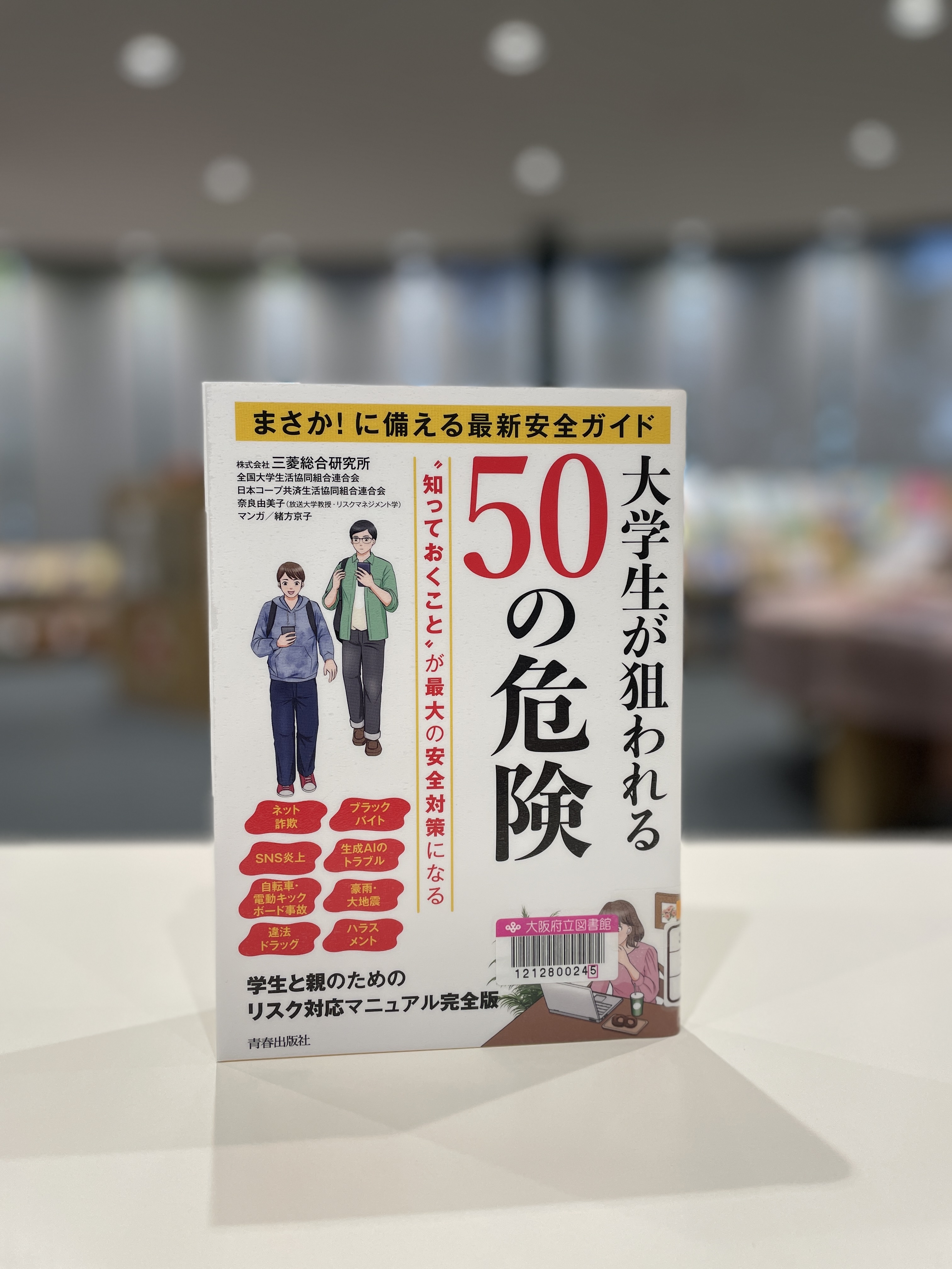 『大学生が狙われる50の危険』の表紙画像。白の背景に赤字でトピックが書かれ、机に座ってパソコンを見る女性とスマホを見ながら歩く男性２人のイラストが描かれている。