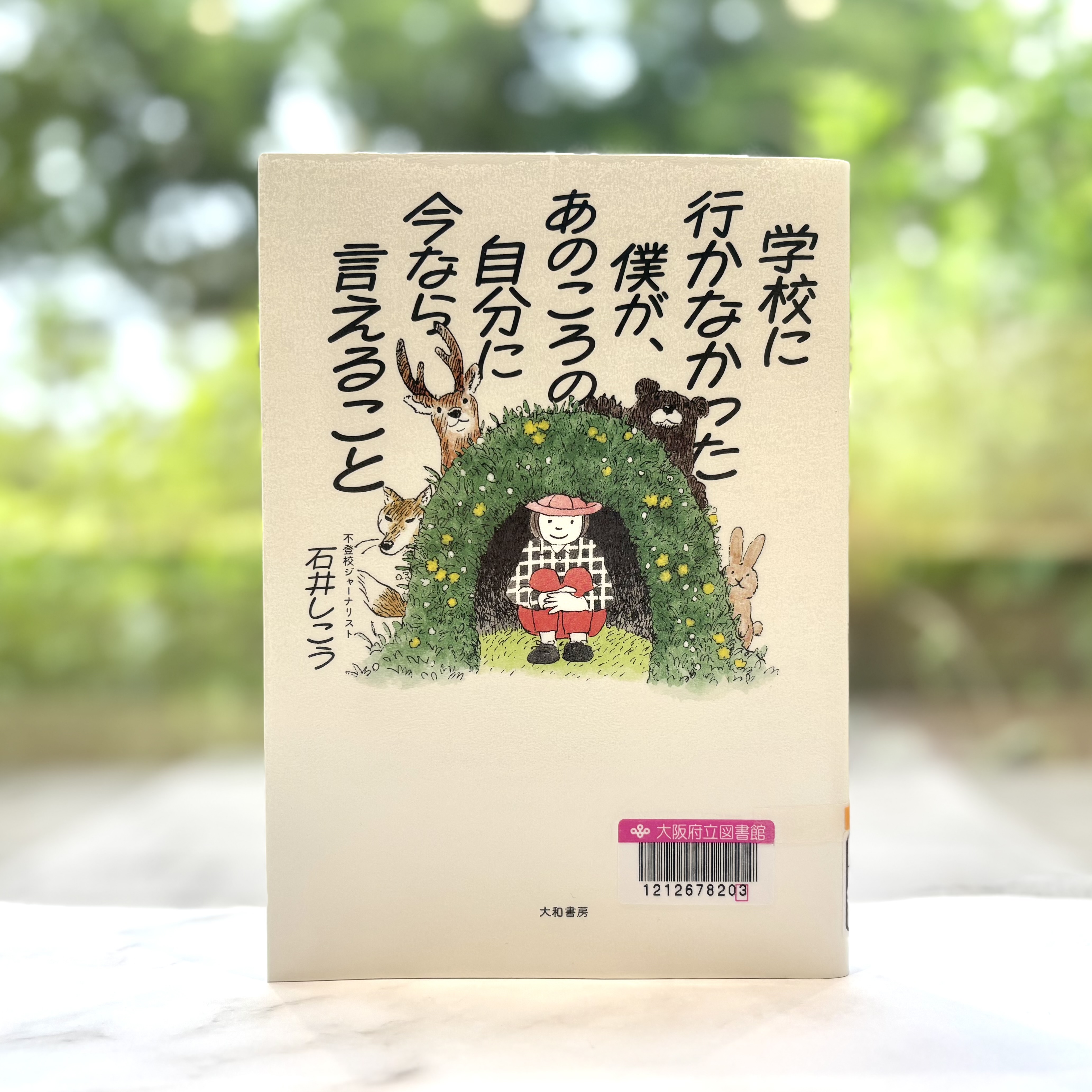 『学校に行かなかった僕が、あのころの自分に今なら言えること』の表紙画像。草木でつくられたほら穴の中にしゃがむ人物とほら穴の外まわりに動物がいるイラスト。タイトルがほら穴や動物たちにかぶさるように、上の空白部分全体に縦書きで書かれている。