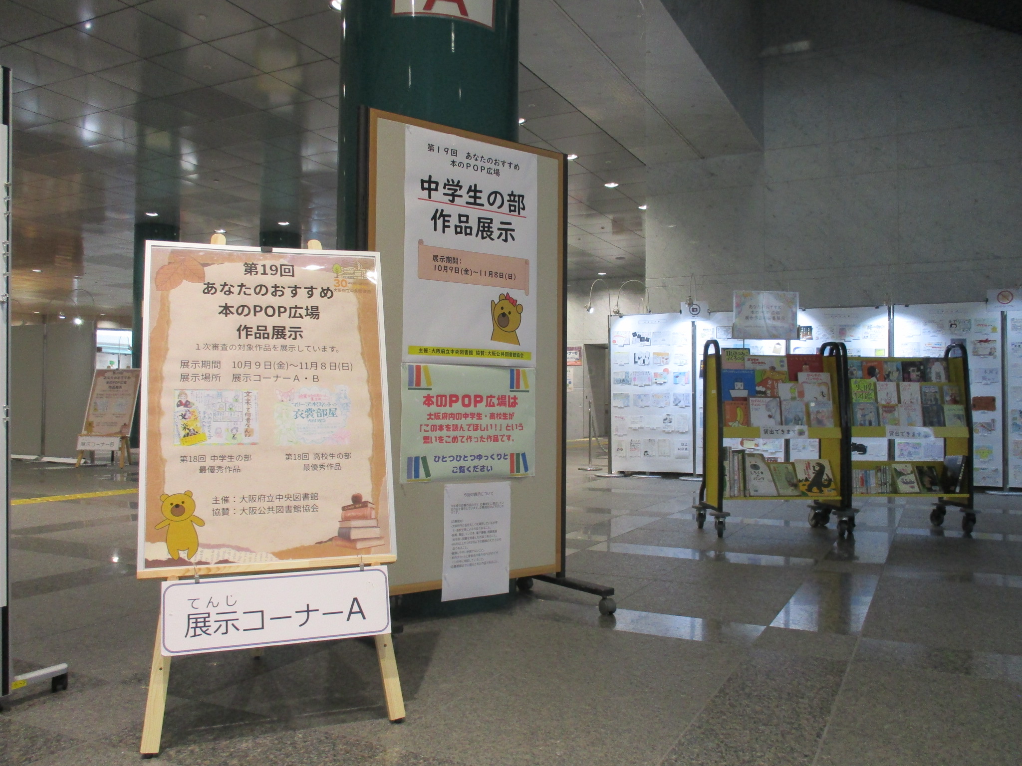 展示コーナーＡの様子―：手前左手に「第19回あなたのおすすめ本のPOP広場　作品展示」と書かれたポスターをのせたイーゼルと「中学生の部　作品展示」と書かれたポスターが貼られた展示ボードが並んでいる。その奥には本を積んだブックトラックが２台並んでいる。さらにその奥にはPOPが貼られた展示ボードが並んでいる。