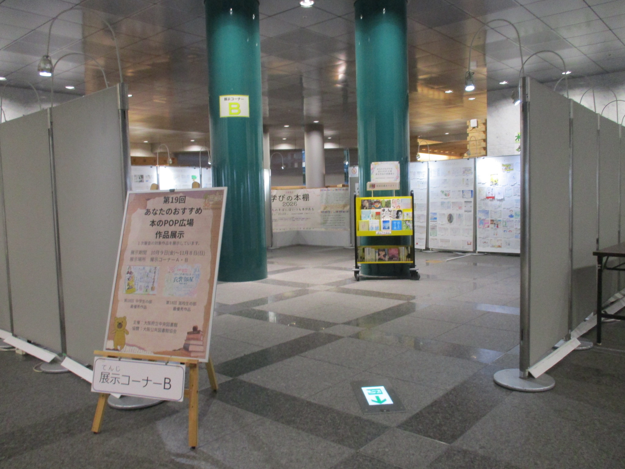 展示コーナーＢの様子―：展示ボードで囲まれた展示スペースの入口左側に「第19回あなたのおすすめ本のPOP広場　作品展示」と書かれたポスターを載せたイーゼルが置かれている。展示スペースの中央の柱前には本を積んだブックトラックが１台置かれている。展示スペースを囲む展示ボードの内側にはPOPが貼られている。