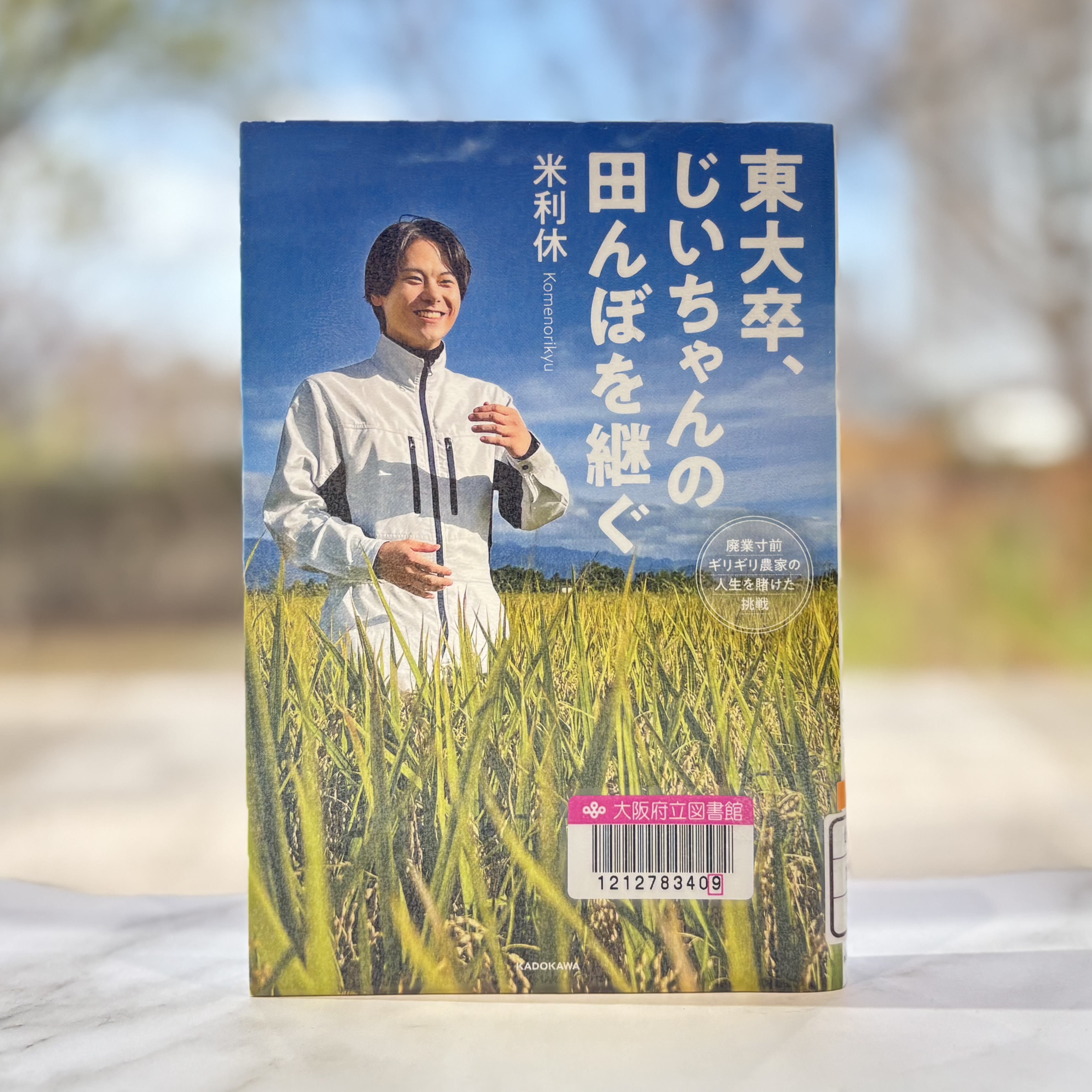大卒、じいちゃんの田んぼを継ぐ：廃業寸前ギリギリ農家の人生を賭けた挑戦』の表紙画像。青空と黄金色の田んぼを背景に、白い作業服を着て立つ男性の写真。