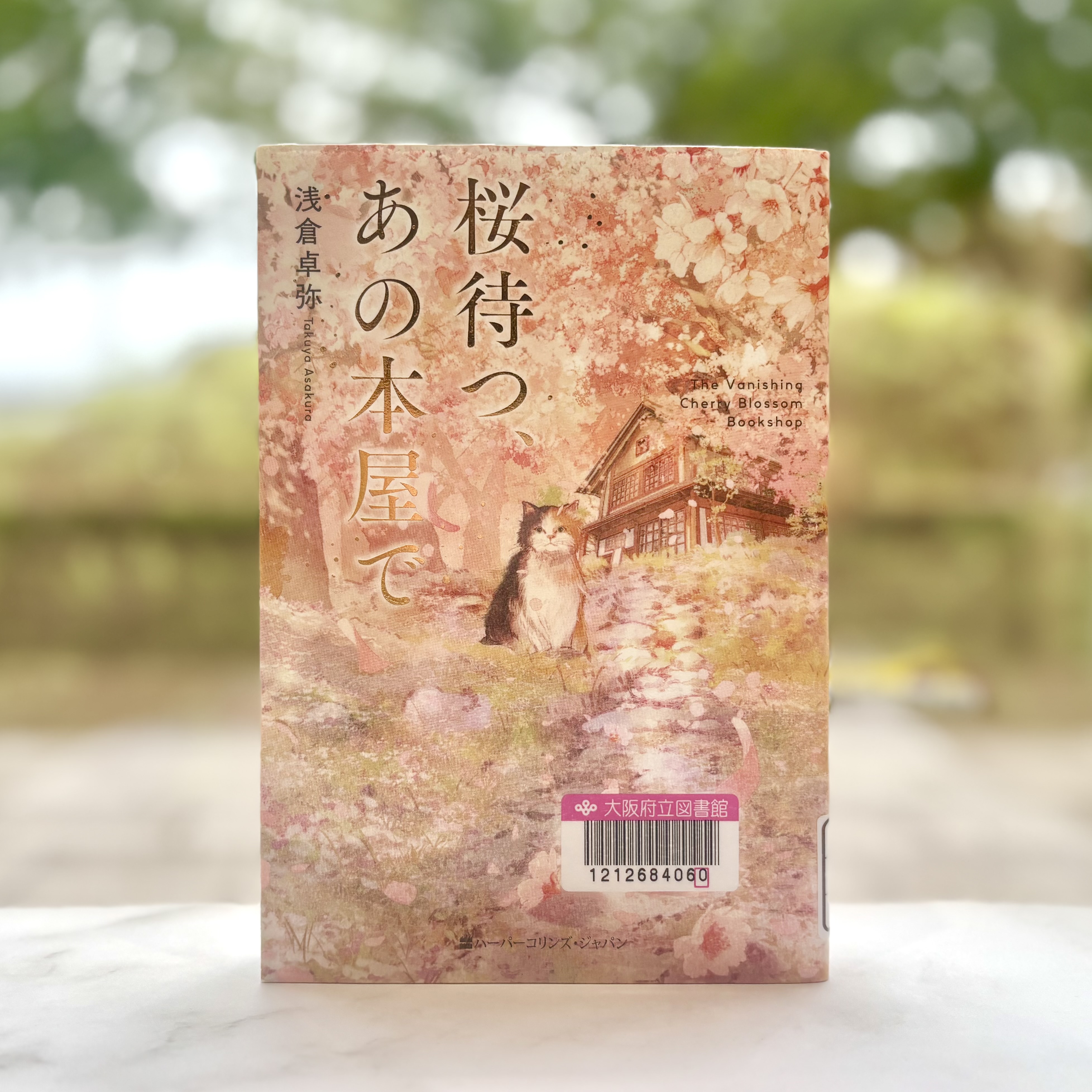 『桜待つ、あの本屋で　The Vanishing Cherry Blossom Bookshop』の表紙画像。和風の家が背後にあり、桜の花びらが舞う桜並木のなか、道のわきに座る猫のイラスト。