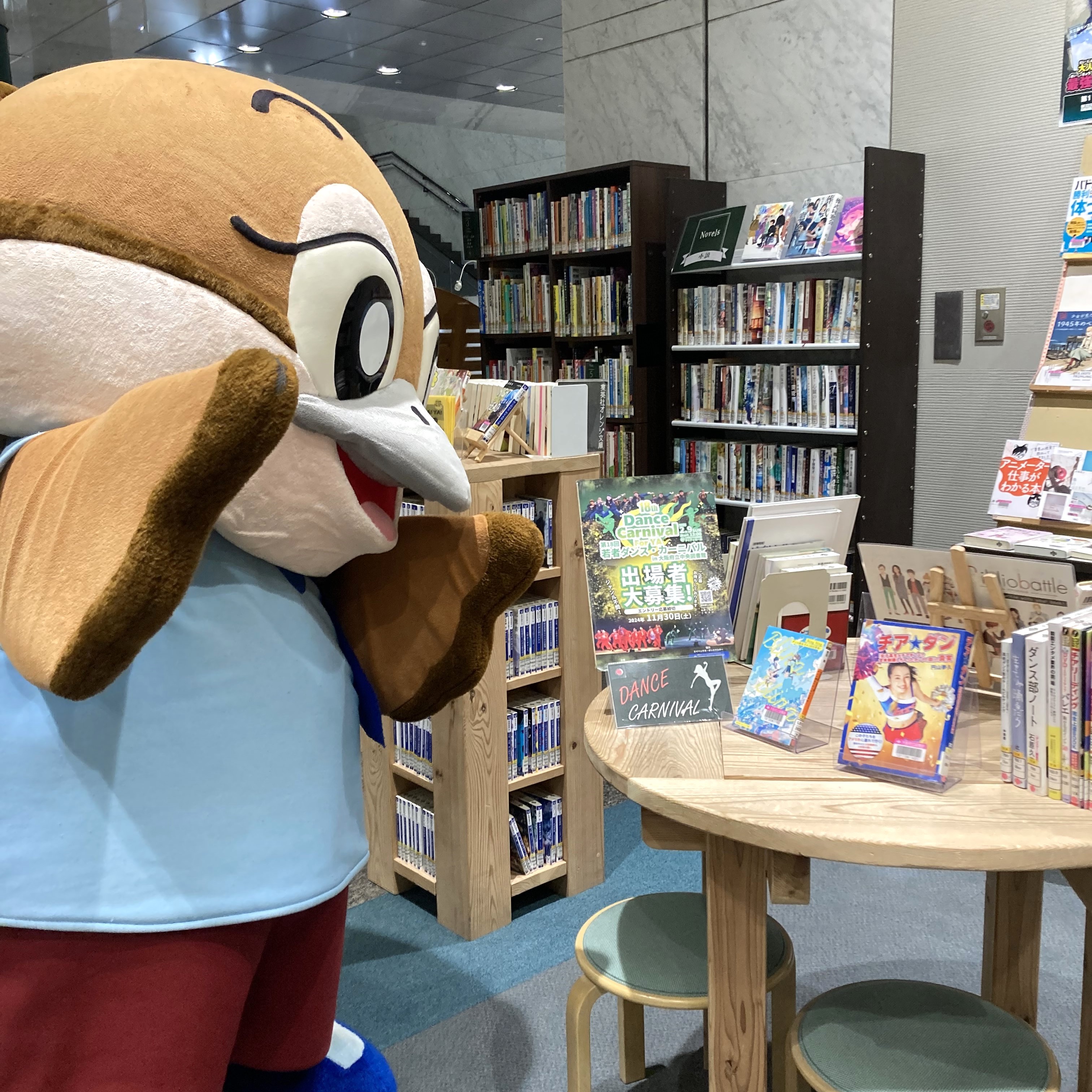 代替テキスト：大阪府のゆるキャラもずやんがYA展示コーナーに来た様子。机の上に並んだ本を眺めている。