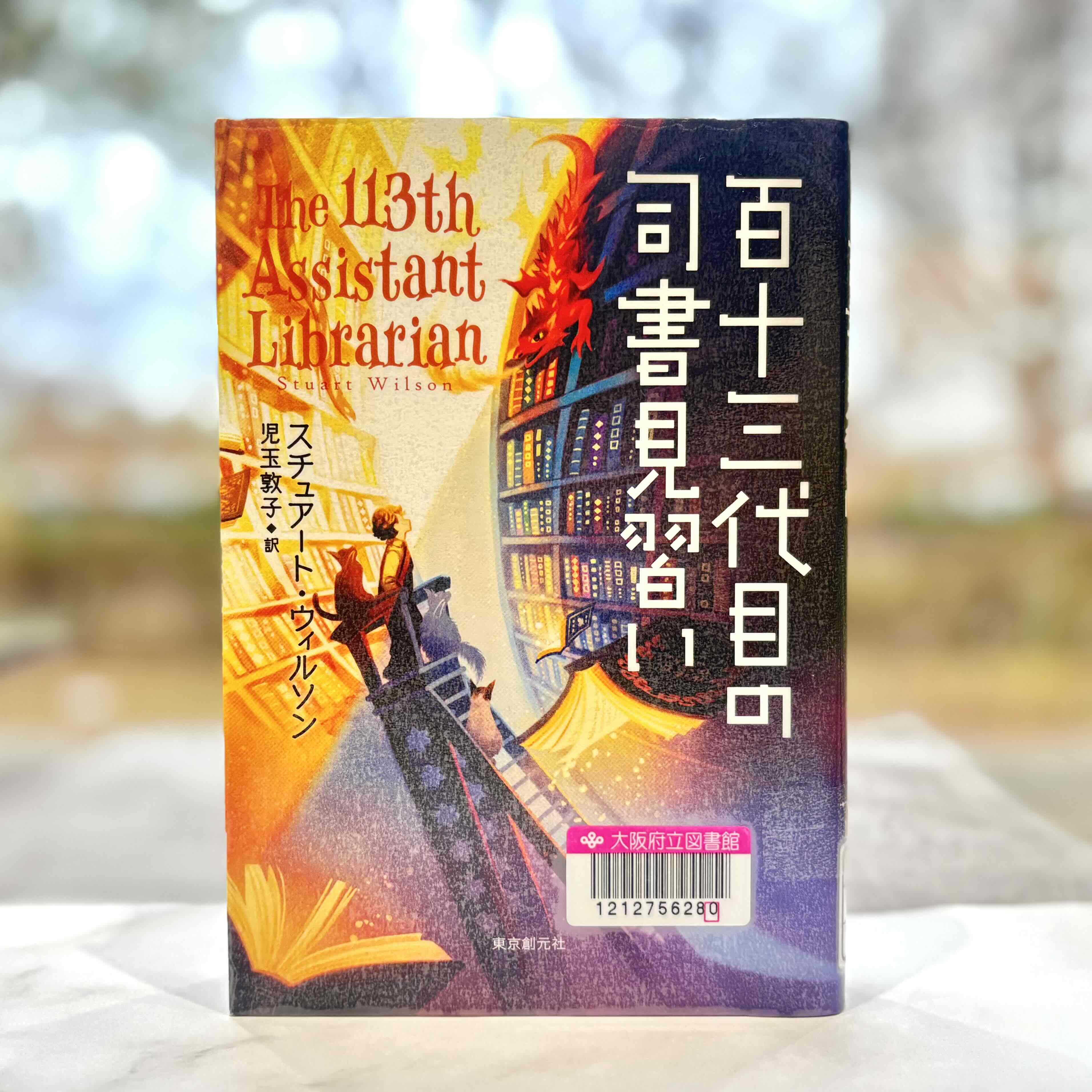『百十三代目の司書見習い』の表紙画像。たくさんの本が並べられた図書館の本棚にいるドラゴンを眺める主人公の男の子と猫のイラスト。